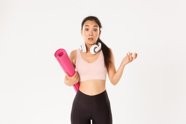   Confused and shocked asian fitness girl, cant understand why gym closed, holding rubber mat and looking surprised at camera,  