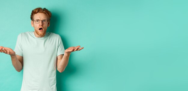 Confused and shocked guy with red messy hair and glasses shrugging and raising hands staring at some