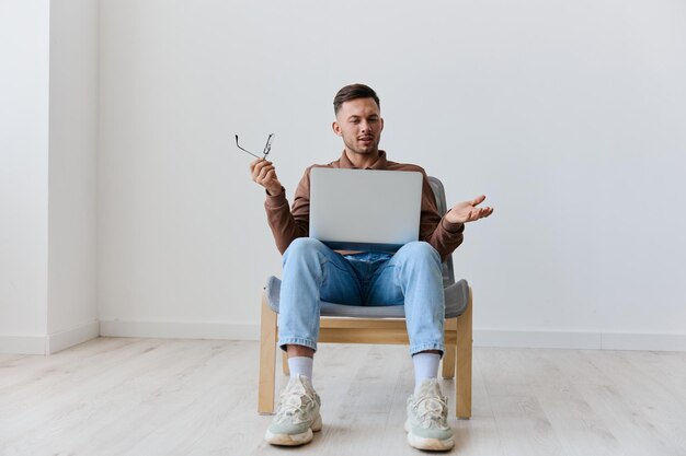 Confused shocked irritated young tanned man with laptop on\
knees can not understand work task computer error say what sinning\
on chair at white home wall shocked by gadget breakdown offer copy\
space