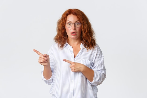 Confused and shocked middle-aged redhead woman in glasses react to something strange, gasping and looking worried while pointing upper left corner, standing white wall.