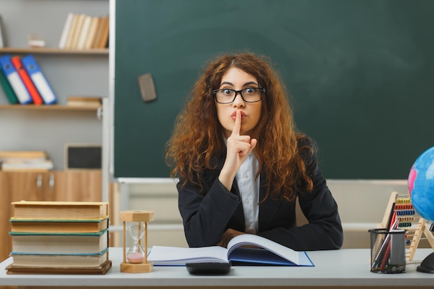 Confuso che mostra gesto di silenzio giovane insegnante femminile che indossa occhiali seduto alla scrivania con strumenti scolastici in classe