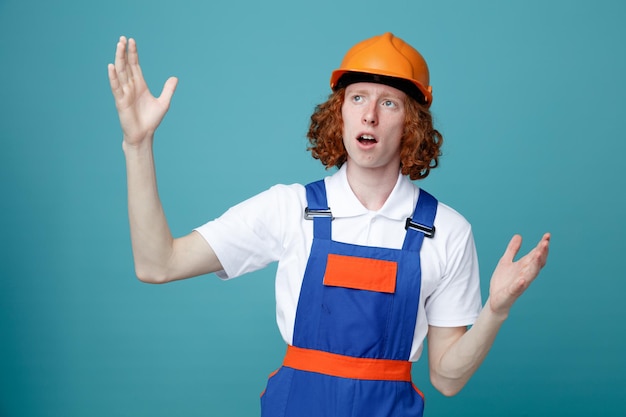 Photo confused showing size young builder man in uniform isolated on blue background