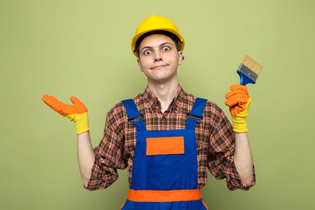 Confused spreading hand young male builder wearing uniform with gloves holding paint brush 