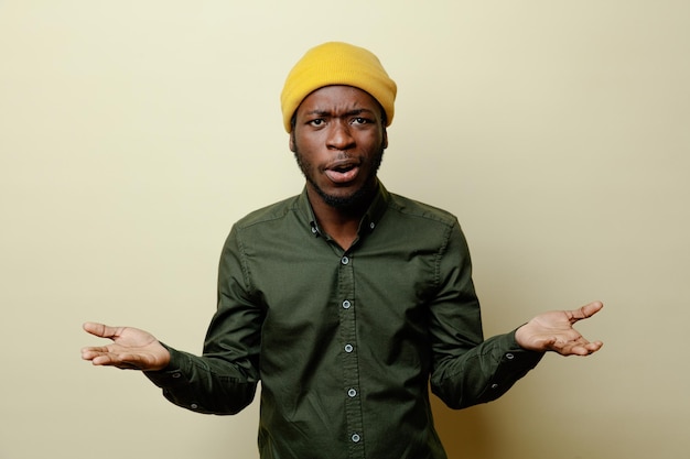Confused spreading hands young african american male in hat wearing green shirt isoloated on white background