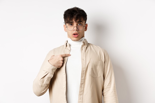 Confused and surprised young man pointing at himself, being chosen or accused, standing on white background