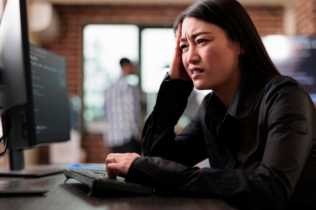 Confused tech engineer sitting doubtful at computer trying to solve network processing errors. Asian computer programmer puzzled by nonfunctional security algorithm and company corrupted database.