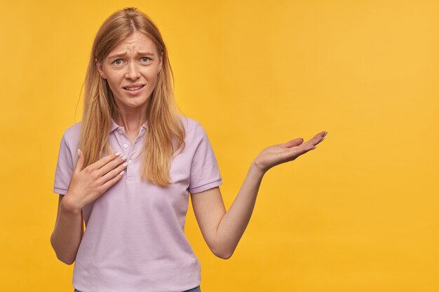 Confused unhappy blonde young woman with freckles in lavender tshirt pointing at herself by hand and holding empty space on palm over yellow wall