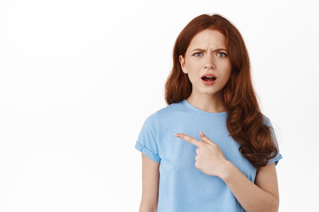 Confused and upset redhead girl, complaining at logo banner, pointing finger left, frowning and grimacing disappointed clueless, standing over white background
