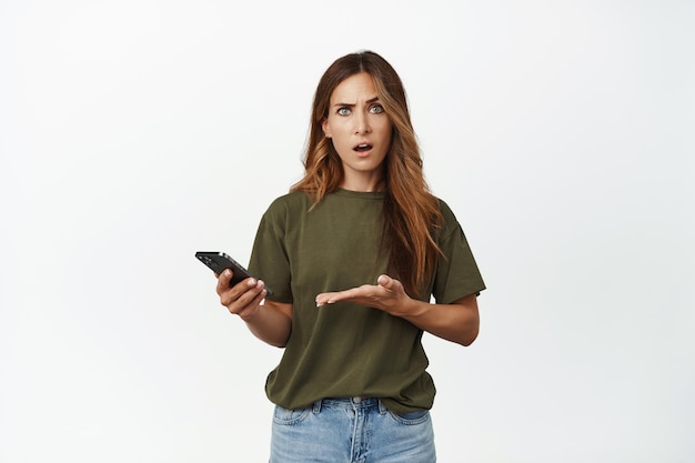 Confused woman pointing at smartphone screen, holding mobile phone and looking bothered at camera, standing against white background. Copy space