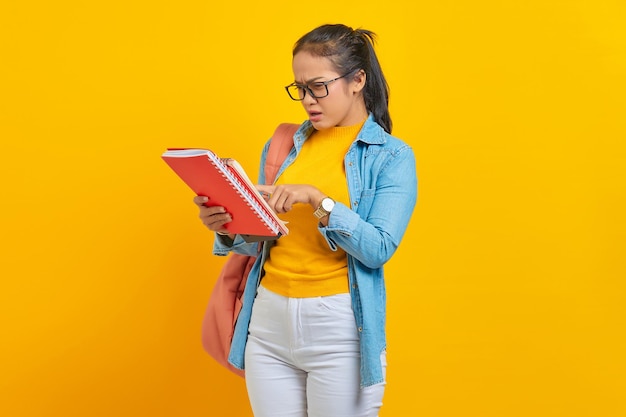 Confused woman student in denim clothes with backpack reading\
in the work notebook isolated on yellow background education in\
high school university college concept