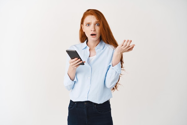 Confused and worried redhead woman receiving bad news on phone looking at camera puzzled and shrugging dont know what happening app crash white background