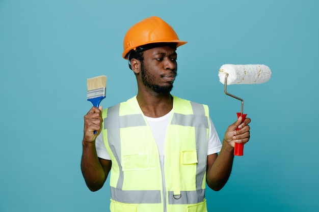 Confused young african american builder in uniform holding roller brush with paint brush isolated on blue background