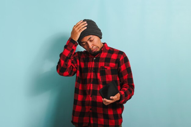 Photo confused young asian man scratching head while looking at his empty wallet isolated on blue backdrop