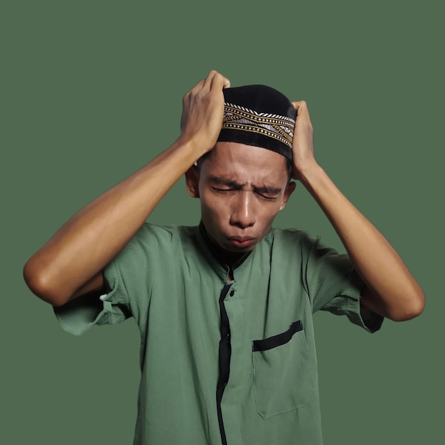 Photo confused young asian muslim man holding head with both hands isolated green background