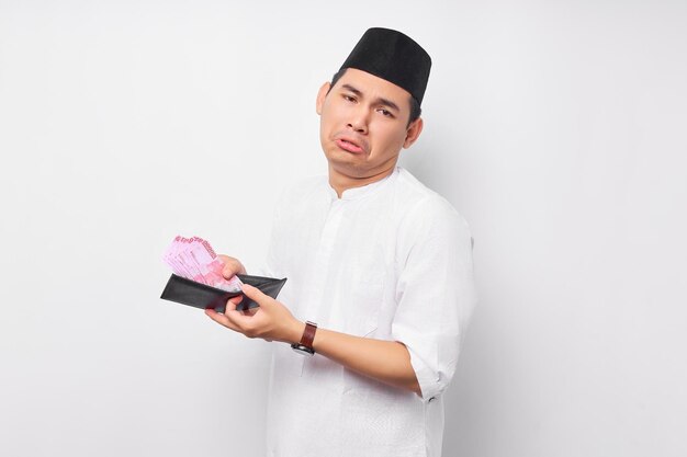 Confused young Asian Muslim man holding wallet of full cash money isolated on white background People religious Islamic lifestyle concept