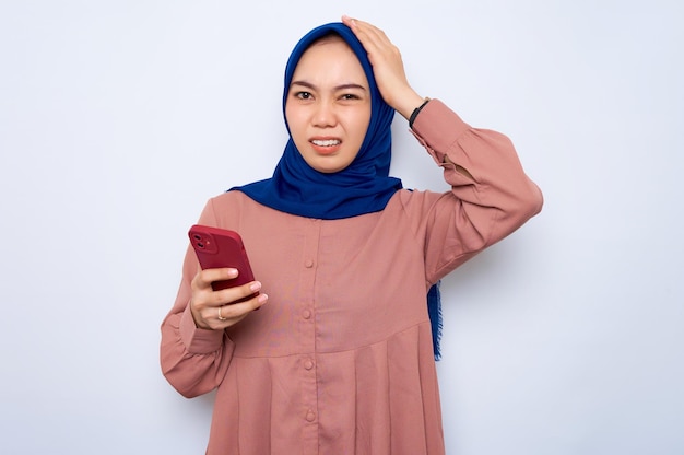 Confused young asian muslim woman in pink shirt holding mobile\
phone isolated over white background people religious lifestyle\
concept