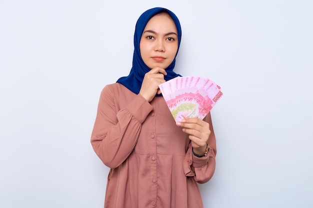 Confused young Asian muslim woman in pink shirt holding money banknotes isolated over white background People religious lifestyle concept