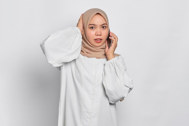 Confused young Asian Muslim woman talking on mobile phone isolated over white background