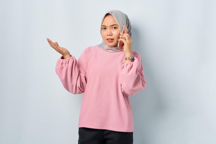 Confused young Asian woman in pink shirt talking on mobile phone isolated over white background