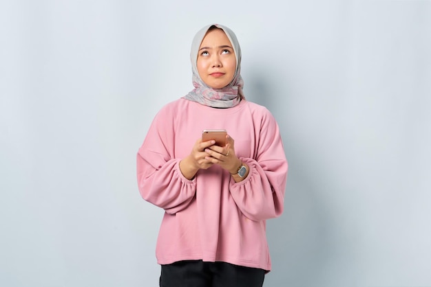 Confused young asian woman in pink shirt using mobile phone and\
looking up isolated over white background