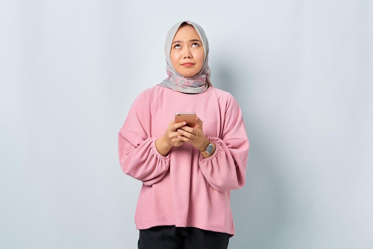 Confused young Asian woman in pink shirt using mobile phone and looking up isolated over white background