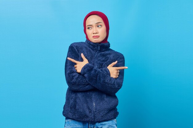 Confused young asian woman pointing sideways and looking\
sideways on blue background