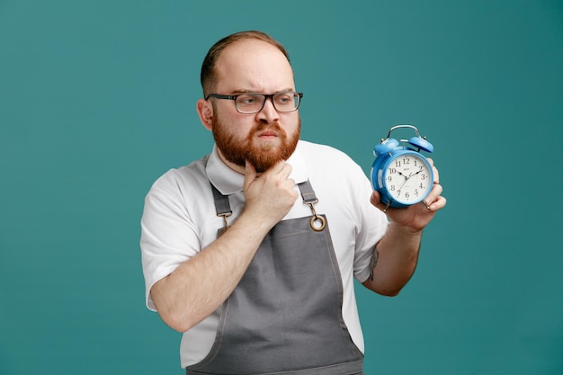 Confused young barber wearing uniform and glasses showing alarm clock keeping hand on chin looking at side isolated on blue background