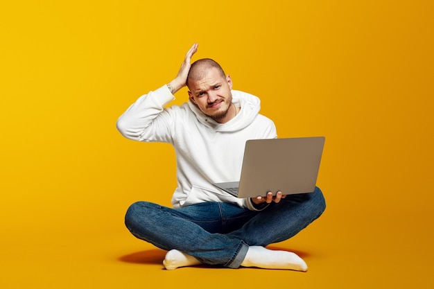 Confused young bearded man sitting on floor holding laptop and scratching his head upset