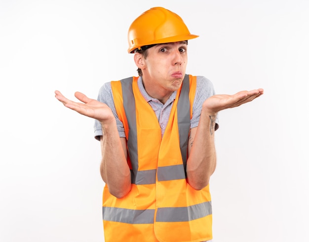 Confuso giovane costruttore in uniforme che spande le mani isolate sul muro bianco