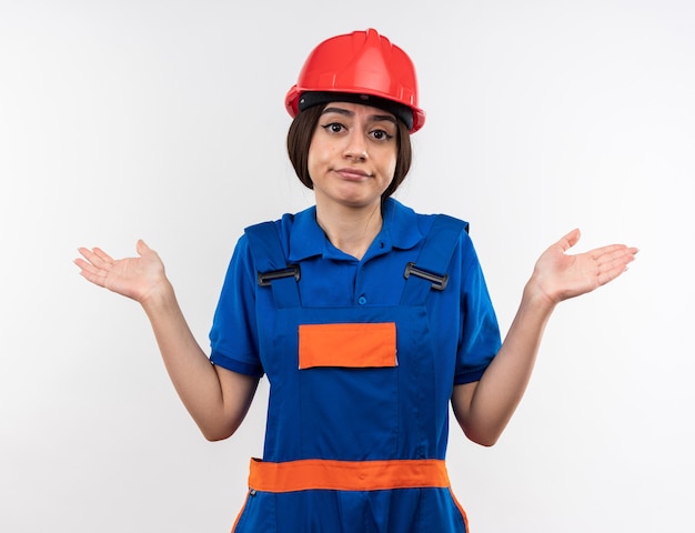 Confused  young builder woman in uniform spreading hands isolated on white wall