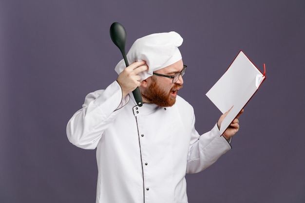 Giovane chef confuso che indossa occhiali uniformi e cappuccio con cucchiaio solido e blocco note guardando il blocco note toccando la testa isolata su sfondo viola