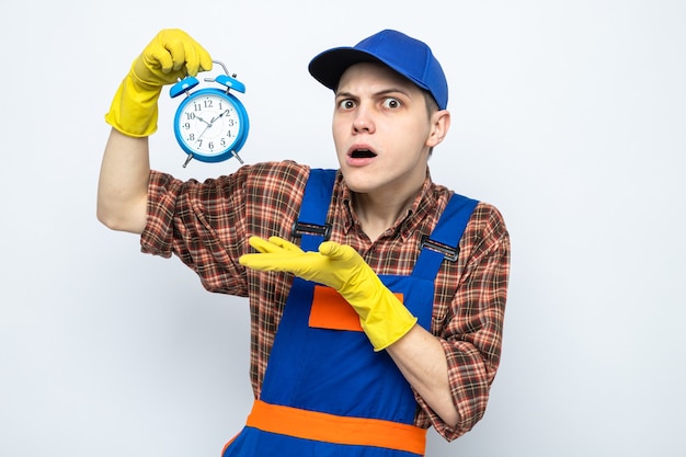 Confused young cleaning guy wearing uniform and cap with gloves holding and points with hand at alarm clock 