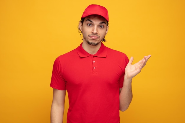 Confused young delivery man wearing uniform and cap looking at camera showing empty hand isolated on yellow background