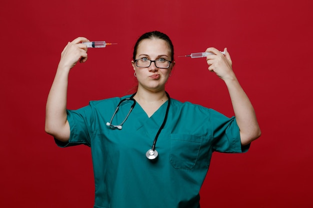 Giovane dottoressa confusa che indossa l'uniforme degli occhiali e lo stetoscopio intorno al collo guardando la fotocamera che tiene le siringhe puntandole alla testa isolata su sfondo rosso