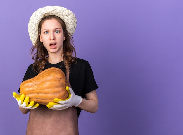 Confused young female gardener wearing gardening hat with gloves holding pumpkin 