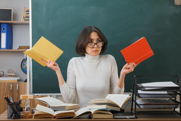 Giovane insegnante confusa che indossa occhiali con in mano libri seduti alla scrivania con strumenti scolastici in classe