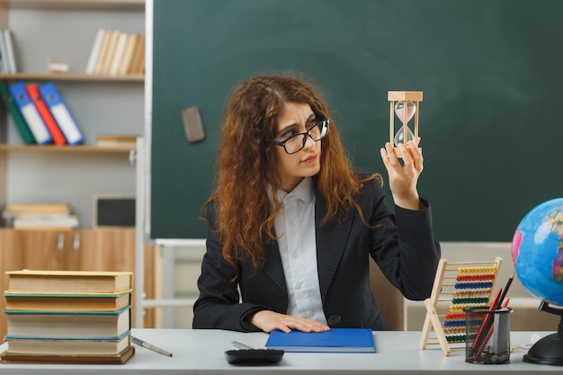 Foto giovane insegnante confusa con gli occhiali che tiene e guarda l'orologio della sabbia seduto alla scrivania con gli strumenti della scuola in classe