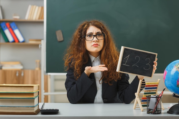 confused young female teacher wearing glasses holding and points at mini chalkboard sitting at desk with school tools in classroom