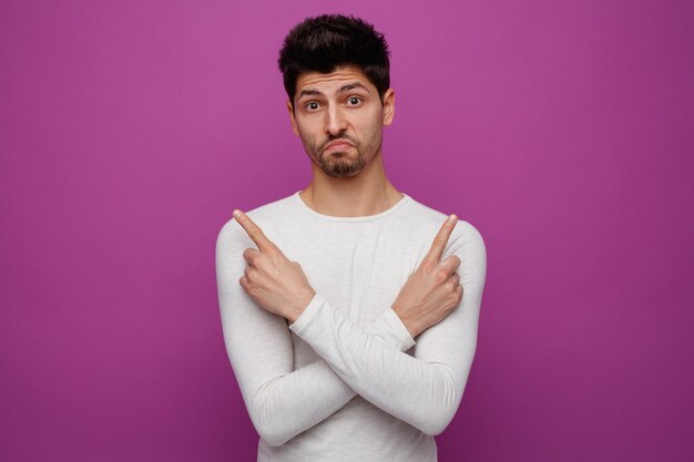 Confused young handsome man looking at camera pointing up with crossed arms on purple background