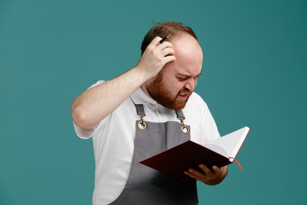 Confused young male barber wearing white shirt and barber apron keeping hand on head holding note pad and pen looking at note pad isolated on blue background