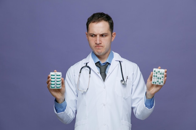 Смущенный молодой врач-мужчина в медицинском халате и стетоскопе на шее показывает упаковки таблеток, глядя на одну изолированную на фиолетовом фоне