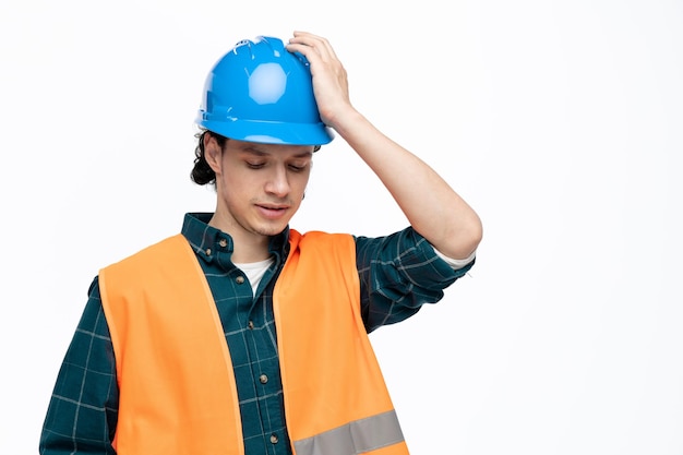 Confused young male engineer wearing safety helmet and safety vest touching his helmet looking down isolated on white background