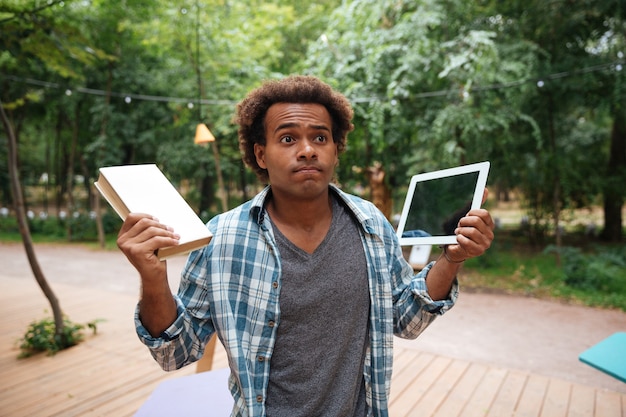 Photo confused young man holding book and tablet outdoors