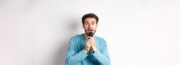 Confused young man looking nervously at camera while singing karaoke holding microphone standing ove