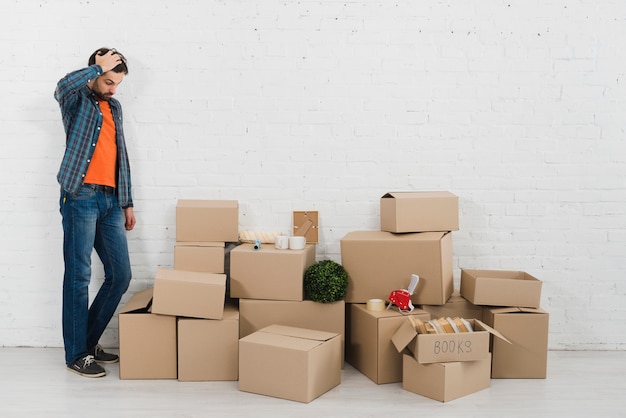 Confused young man looking at piles of cardboard boxes against white brick wall