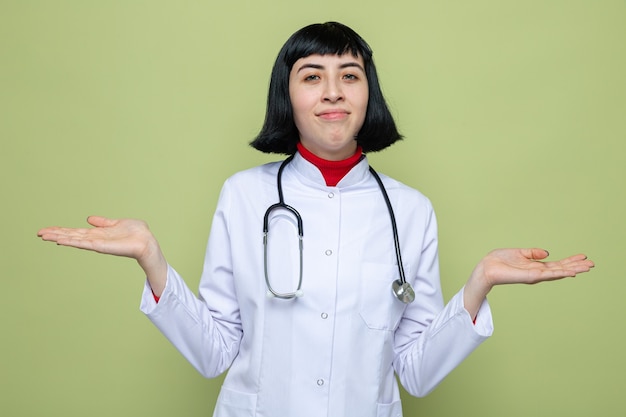 Confused young pretty caucasian girl in doctor uniform with stethoscope holding hands open