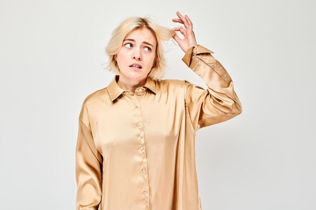 Confused young woman in beige blouse holding hair strand with dry ends isolated on grey background