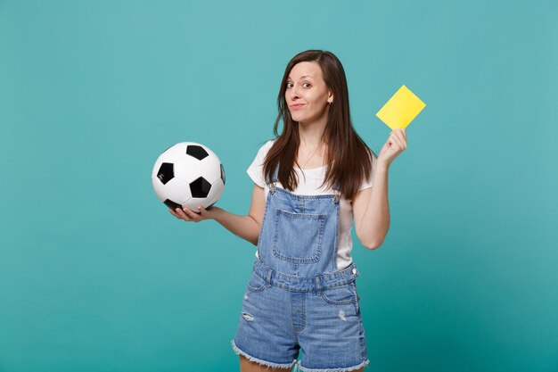 Photo confused young woman football fan support team with soccer ball, yellow card propose player retire from field isolated on blue wall turquoise background. people emotions, sport family leisure concept.