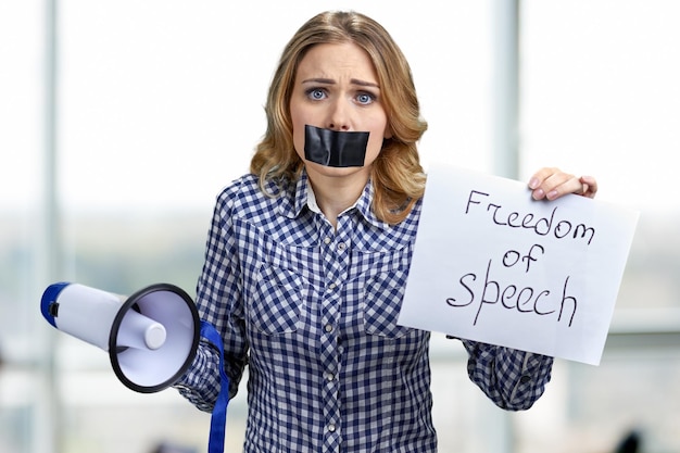 Confused young woman holding card with inscription Freedom of speech. Blur interior background.