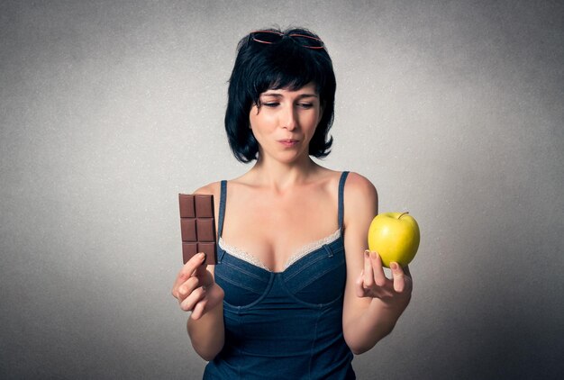 Foto giovane donna confusa con una barretta di cioccolato e una mela sullo sfondo grigio